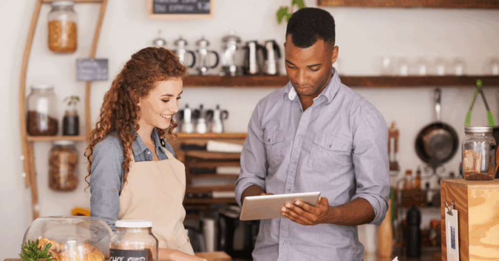 Two cafe employees reviewing business data on a tablet behind a coffee shop counter.