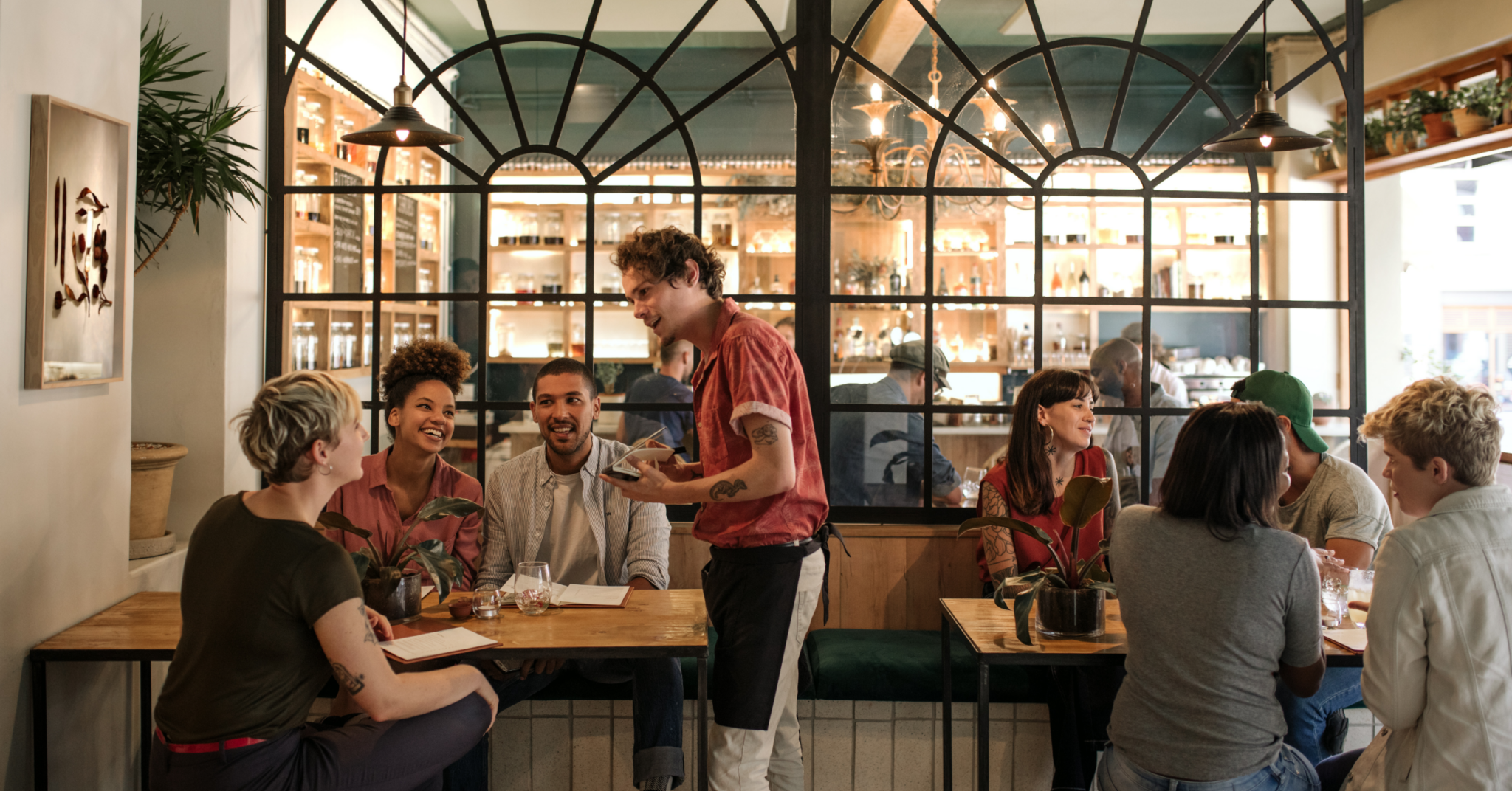 Restaurant server taking an order while customers dine at tables in a busy restaurant dining area.