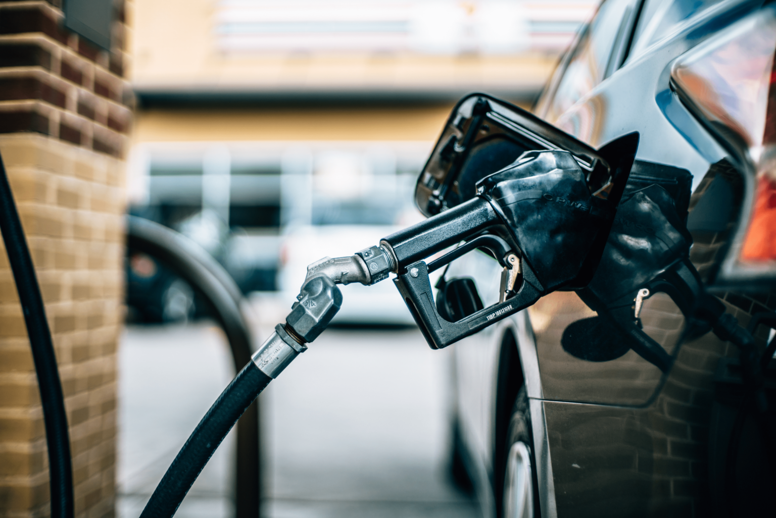 Close-up of a fuel pump nozzle inserted into a vehicle fuel tank, with the gas station forecourt visible in the background.