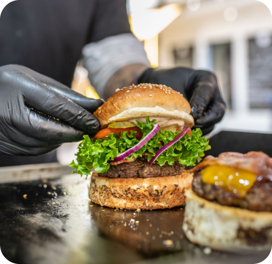 Food service worker wearing black gloves assembling a gourmet burger topped with lettuce, red onion, and sauce on a toasted bun.