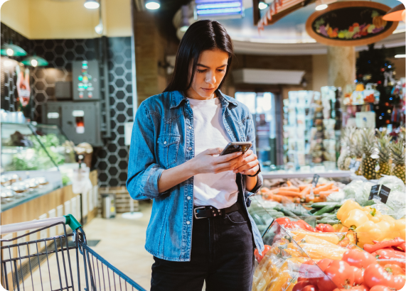 Person standing beside a grocery cart using a smartphone near a fresh produce section stocked with vegetables and packaged foods.
