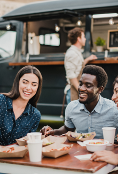 Customers seated at an outdoor table enjoying meals beside a food truck, with serving windows and dining items visible.