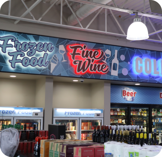 Convenience store beverage section featuring illuminated Frozen Food, Fine Wine, and Beer signage above refrigerated cases and stocked shelves.