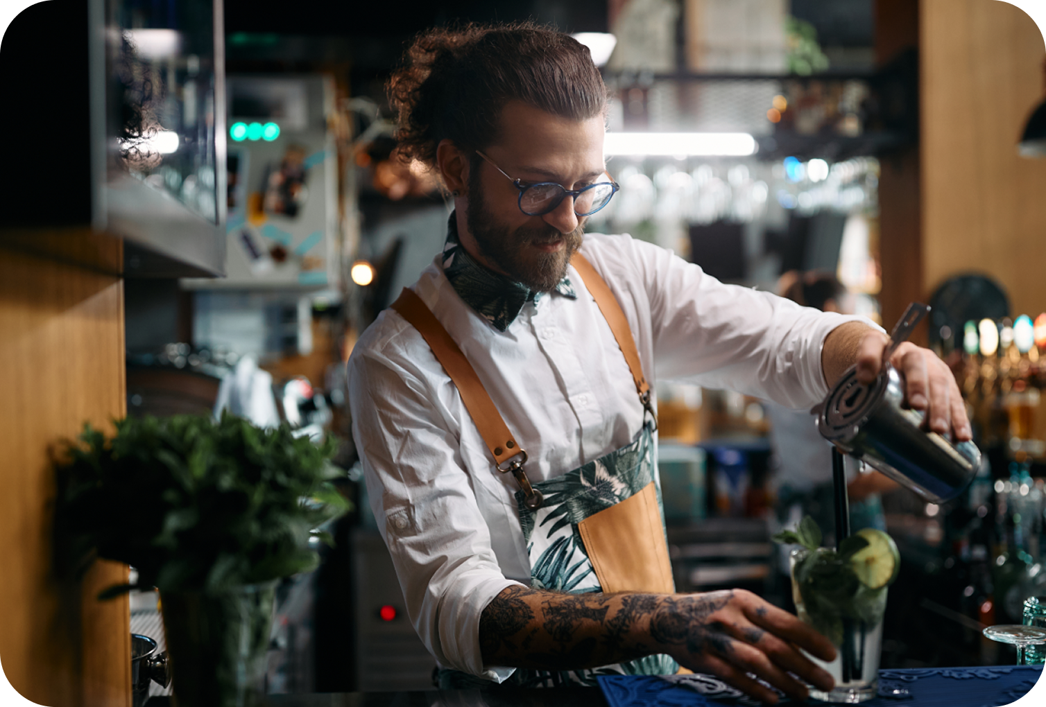 Bar staff member preparing a cocktail behind a bar counter while pouring a beverage into a glass garnished with lime slices.