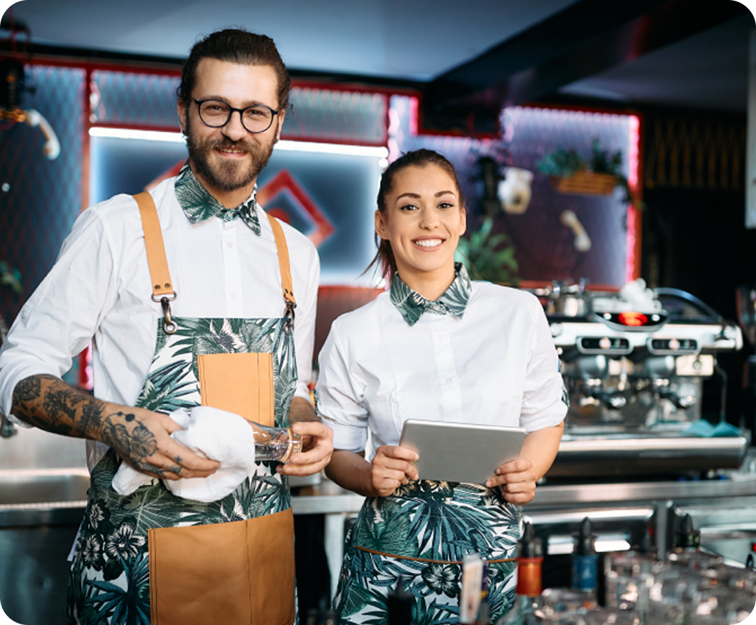Bar staff standing behind a bar counter with a tablet, beverage preparation equipment, and drink service stations in the background.