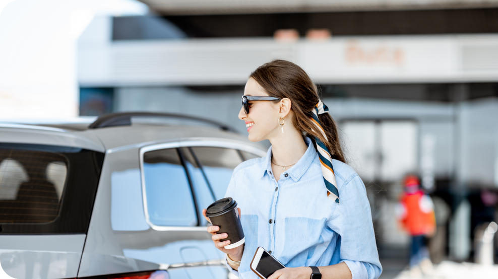 Customer standing beside a vehicle while holding a coffee cup and mobile phone at a fuel station.