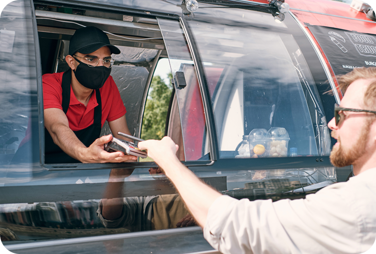 Customer making a contactless payment at a food truck service window using a payment card while a staff member holds a mobile payment terminal.
