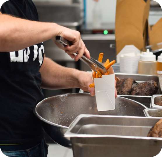Food service worker using tongs to place French fries into a takeout container at a commercial kitchen preparation station.