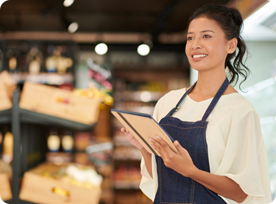 Store employee holding a tablet in a grocery store aisle with produce displays and retail inventory shelves in the background.