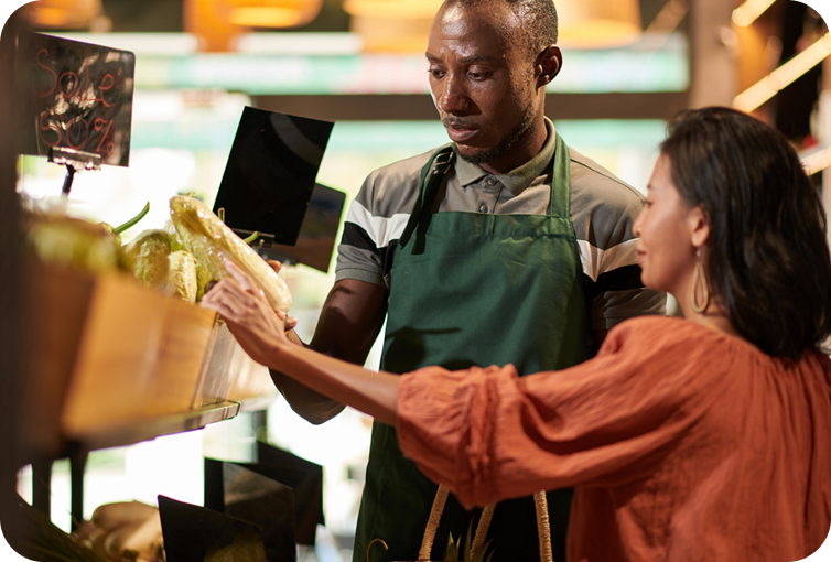 Customer selecting fresh produce from a grocery store display while a store employee assists nearby.