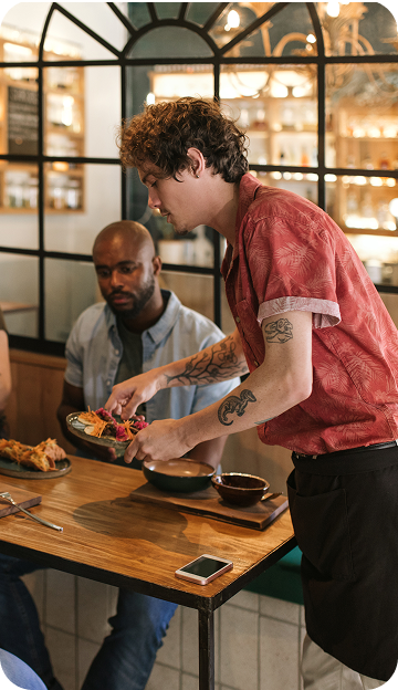 Restaurant staff member serving food to guests seated at a dining table in a restaurant setting.