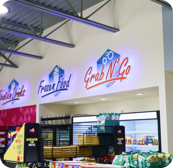 Convenience store interior featuring beverage coolers, product displays, snack aisles, and illuminated store signage.